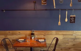 An empty table for 2 against a blue wall with hanging kitchen utensils