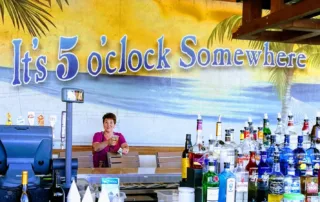 Lady under a "It's five O'clock Somewhere" sign behind a stocked bar.