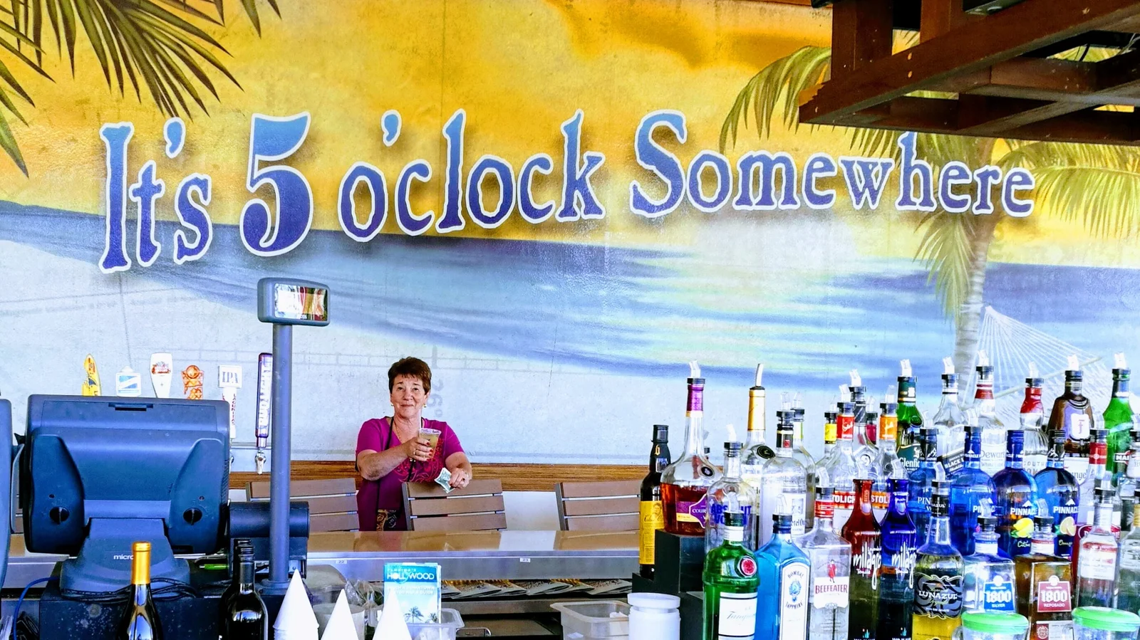 Lady under a "It's five O'clock Somewhere" sign behind a stocked bar.